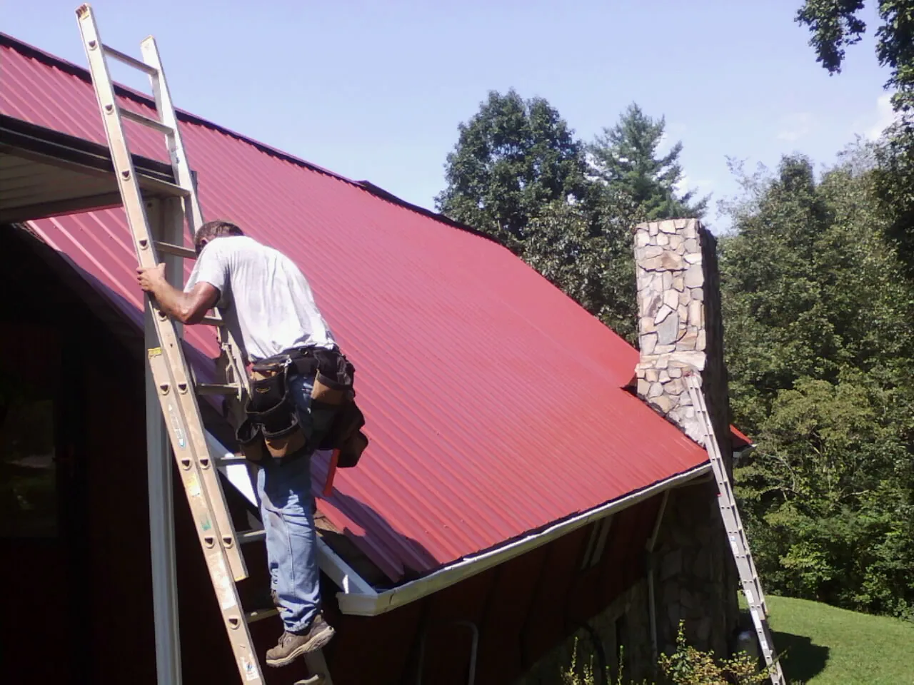 Roofer on a ladder beside a red metal roof and stone chimney — Soffit & Fascia Repair by Roofing Brothers in Hubbardsville
